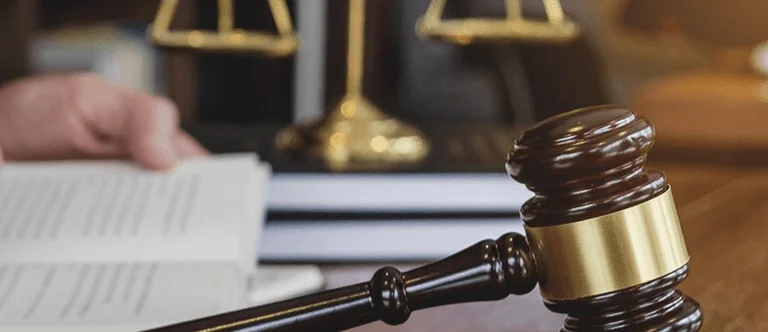 A lawyer's hand writes notes in a book, with a gavel and scales of justice prominently displayed on a desk.