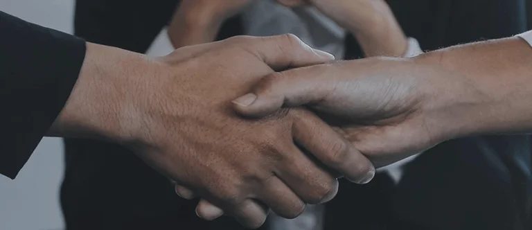 Two hands shaking in a business setting, symbolizing partnership and agreement, with a blurred figure in the background.