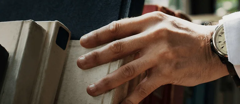 A close-up of a man's hand in a suit, reaching for a book on a shelf filled with various books.