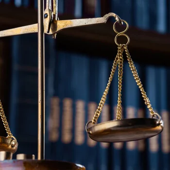 Close-up of a gold balance scale hanging in front of blurred bookshelves, symbolizing justice and law.