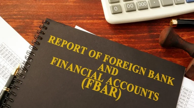 Spiral-bound report titled "Report of Foreign Bank and Financial Accounts (FBAR)" on wooden desk with pen, calculator, and documents.