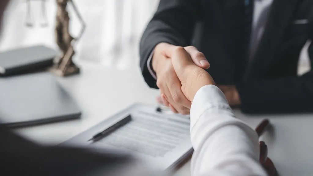 Two professionals shaking hands over a desk with a contract and pen.