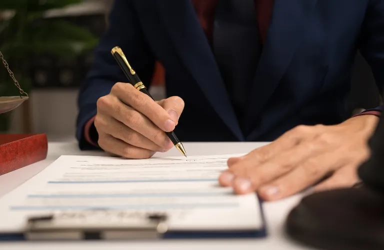 Professional in dark suit signing official document with pen at white desk.