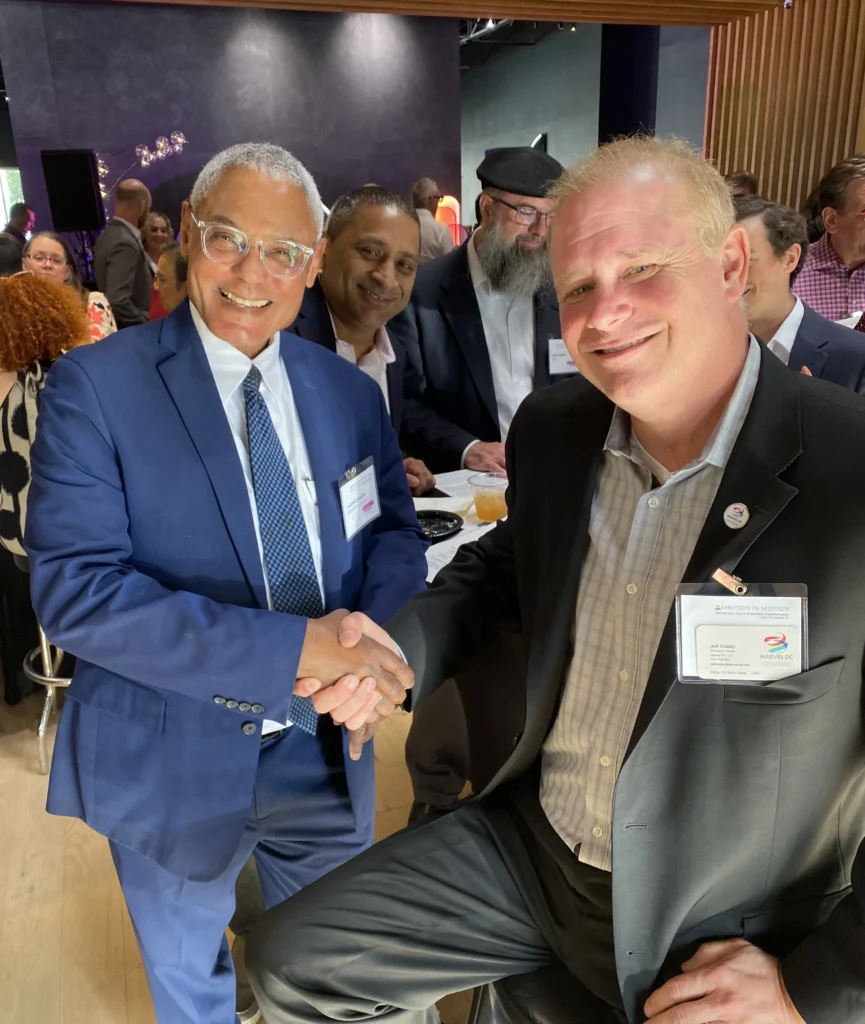 Two professionally dressed men shaking hands and smiling at a networking event.