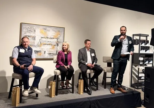 Four professionals on stage; three seated on stools, one standing speaking into a microphone.