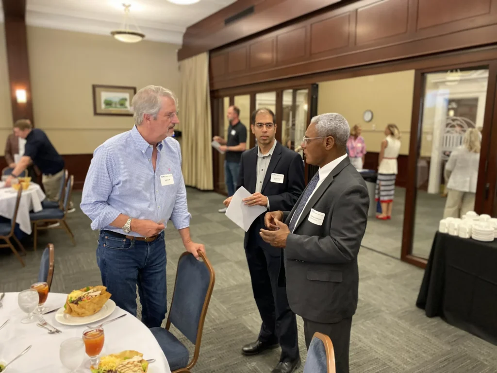 Three men in business attire engaged in conversation in a formal dining room with tables set for a meal.