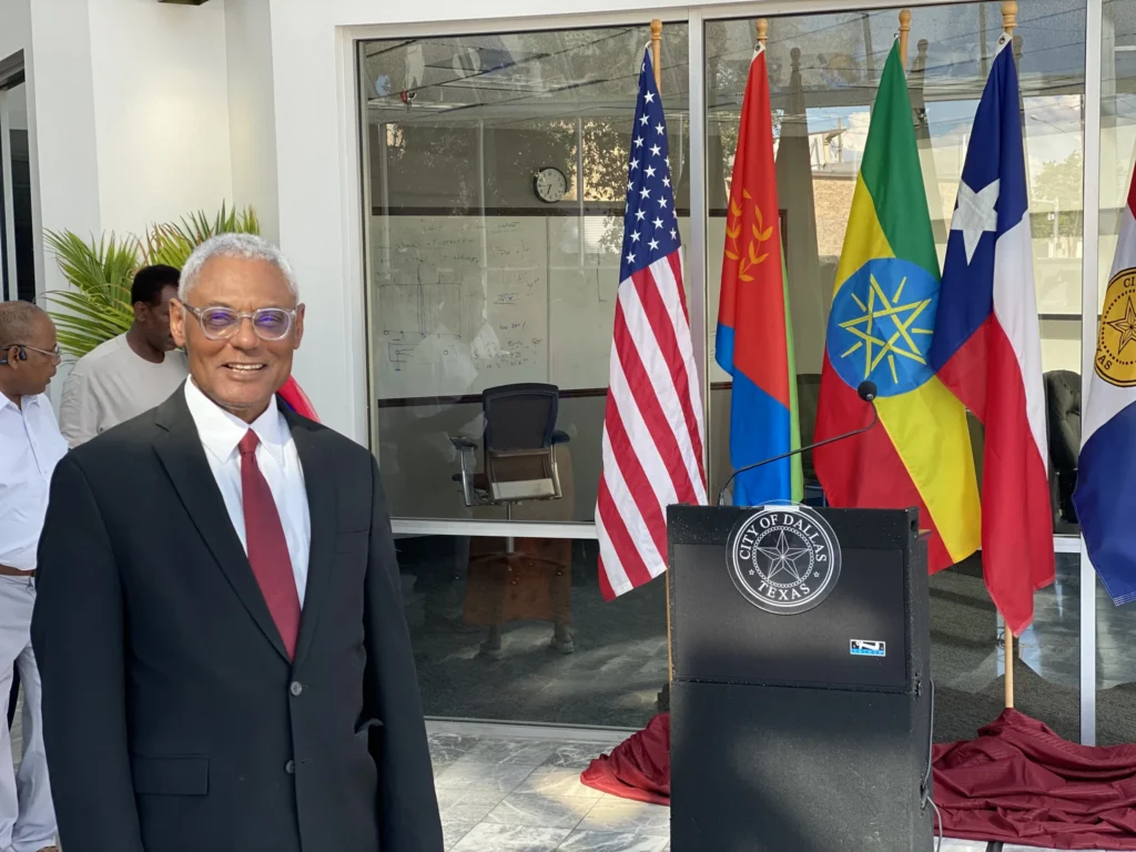 Professional man in black suit and red tie standing near City of Dallas podium with multiple national flags behind.