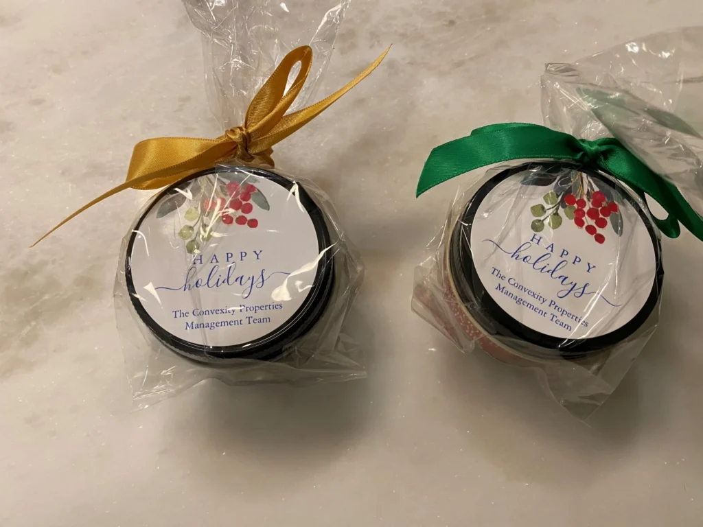Two round holiday gift jars wrapped in clear plastic with gold and green ribbons, labeled "Happy Holidays" from The Convexity Properties Management Team.
