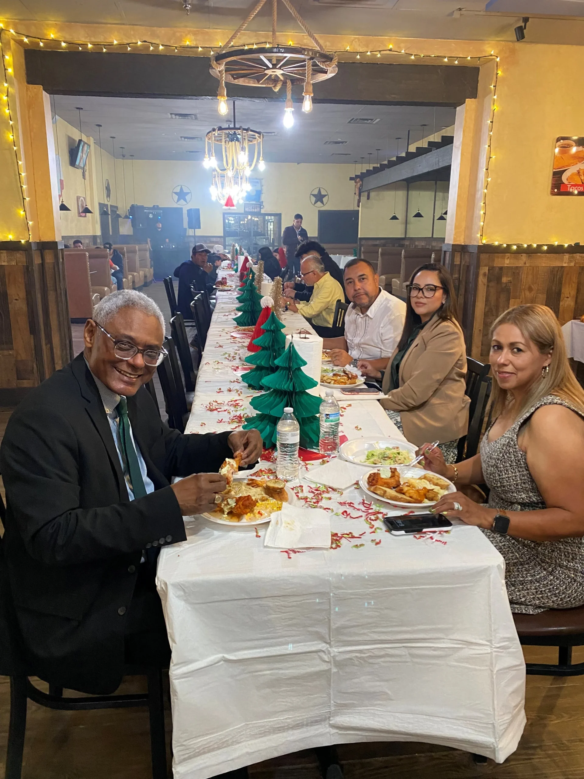 Group of professionally dressed adults seated at a long, decorated dining table with food and water bottles in a warmly lit restaurant.