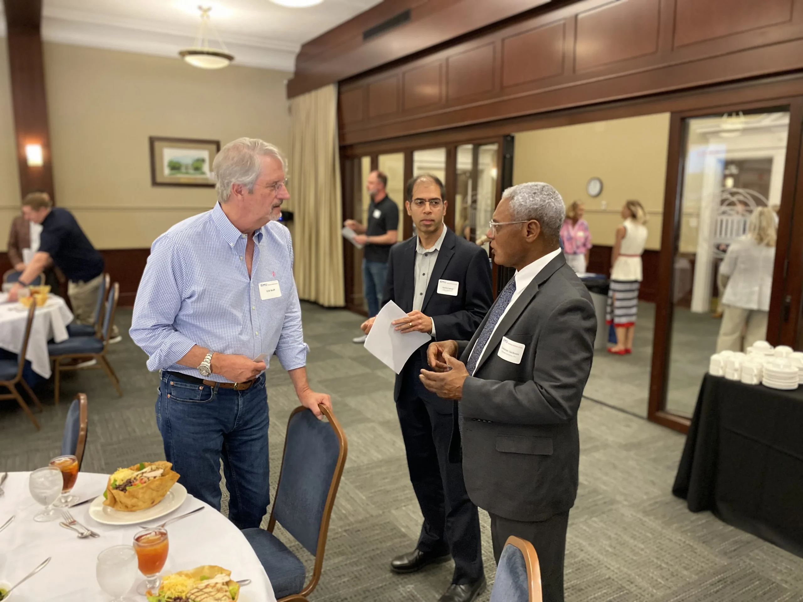 Three professionally dressed men engaged in conversation at a formal indoor event with tables set for dining.