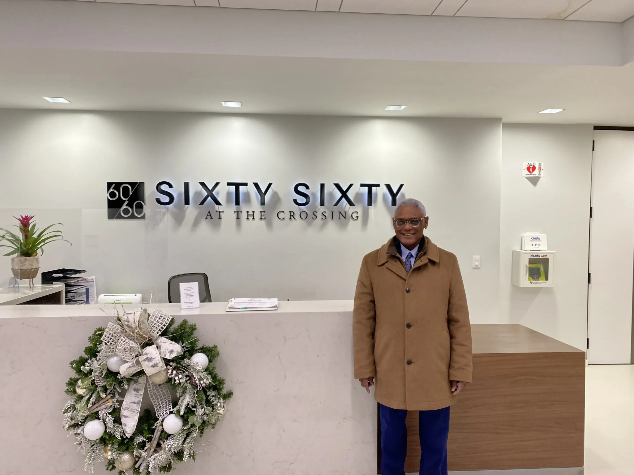 A man in a brown coat and blue pants standing in a modern office lobby with a Christmas wreath on the wall.