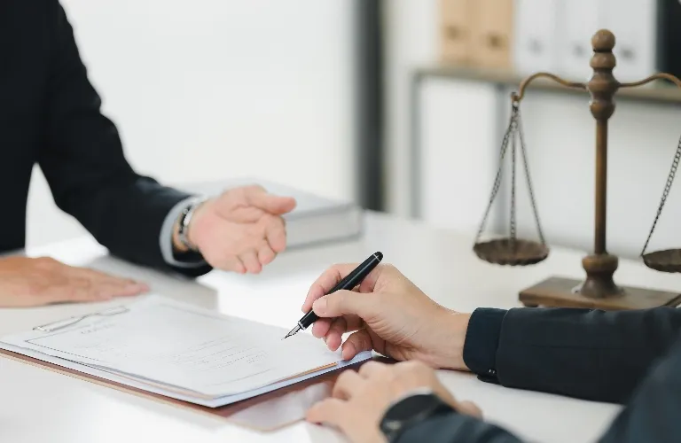 Two individuals in formal attire discussing and signing documents near a brass balance scale on a white desk.