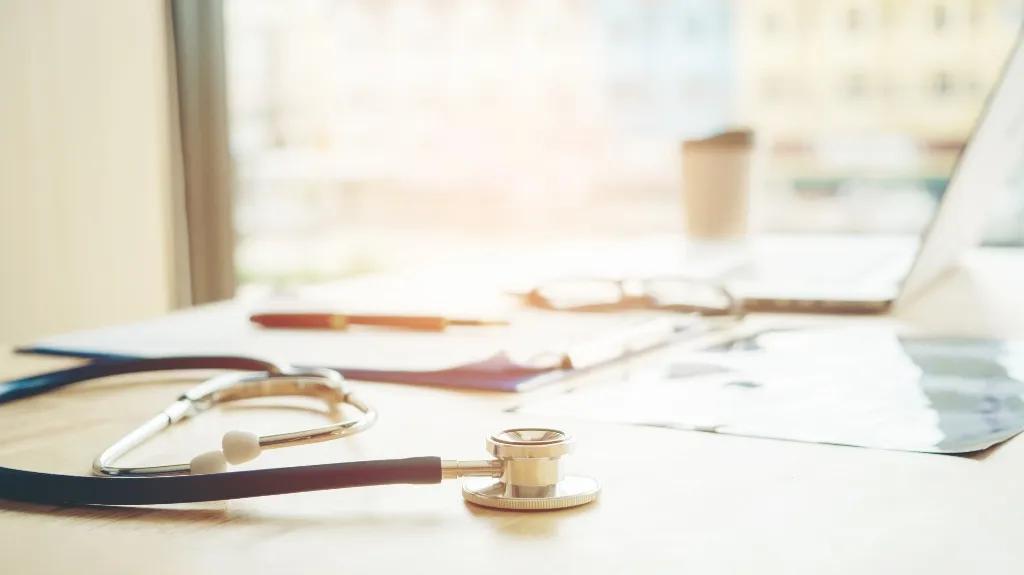 Stethoscope on wooden desk with blurred clipboard, pen, laptop, and coffee cup in background.