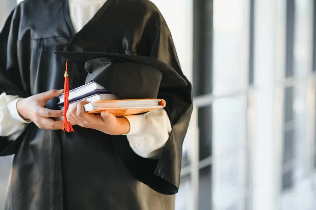 Individual wearing black graduation gown holding books and a graduation cap indoors.