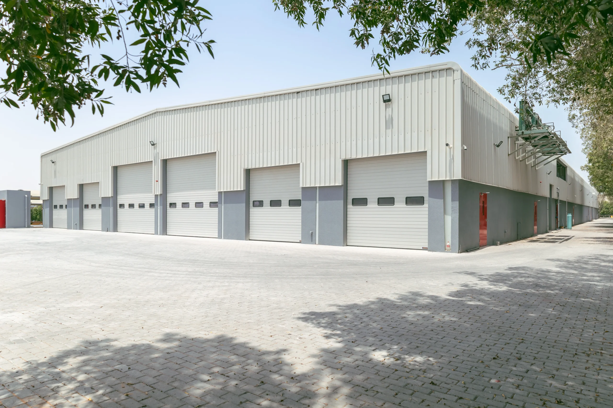 Exterior view of a large industrial warehouse with multiple gray roll-up doors and paved ground.