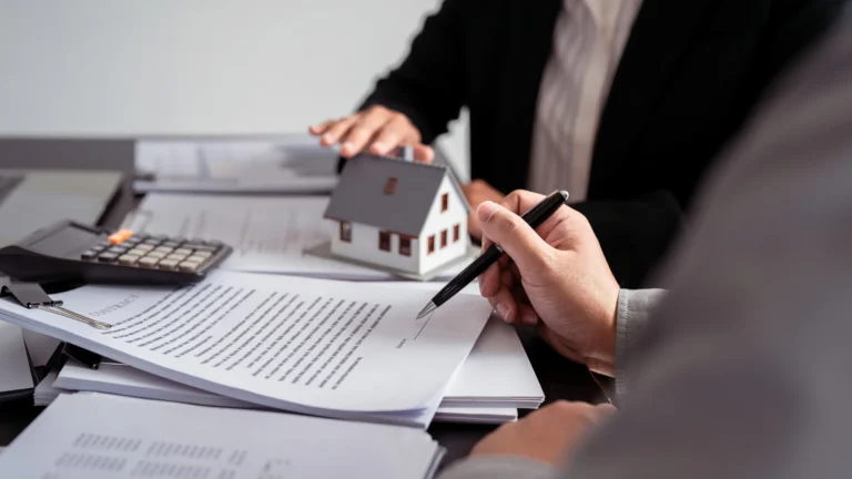 Two professionals reviewing real estate contract documents with a small house model and calculator on the table.
