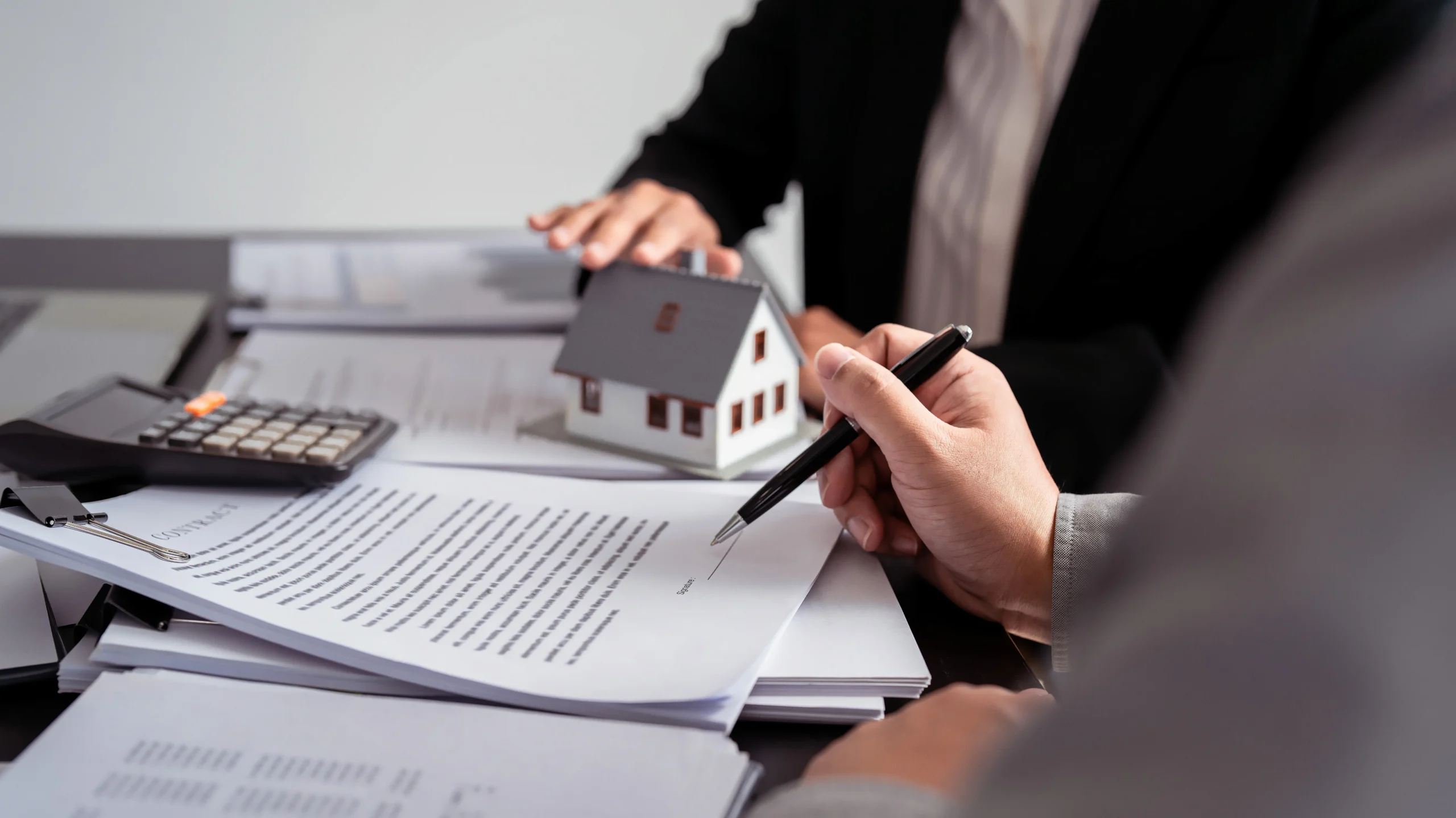 Two professionals reviewing real estate contract documents with a small house model and calculator on the table.