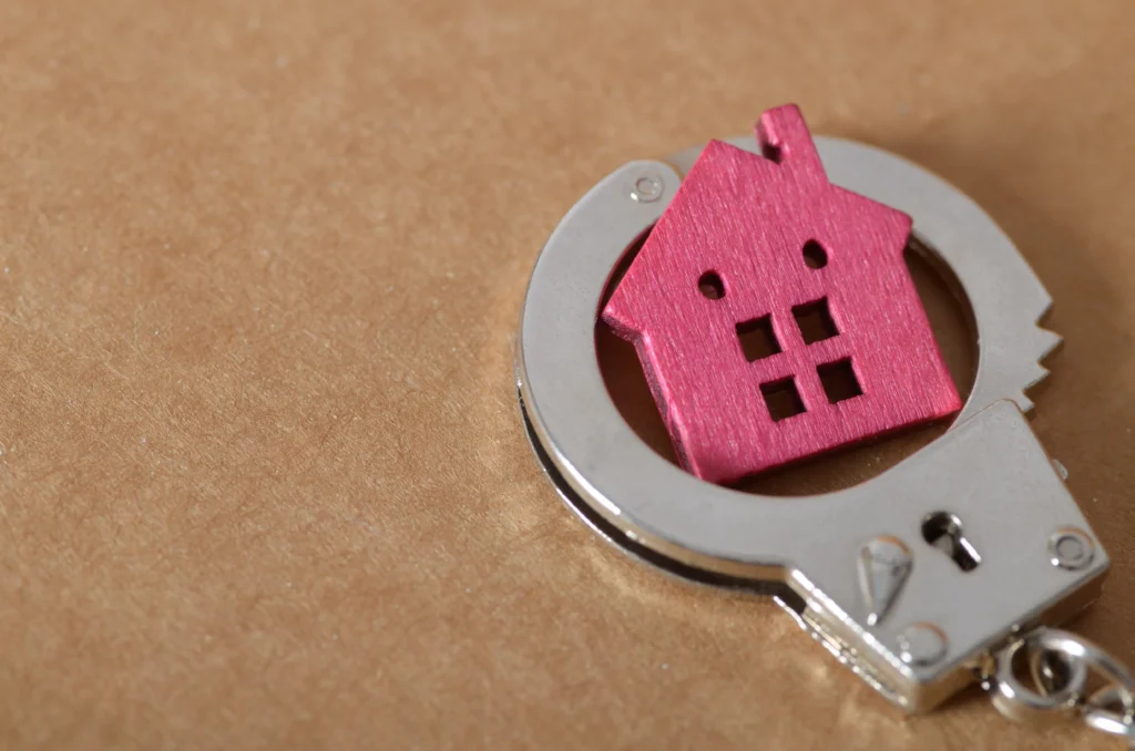 Silver handcuff encircling a small pink wooden house on brown surface.