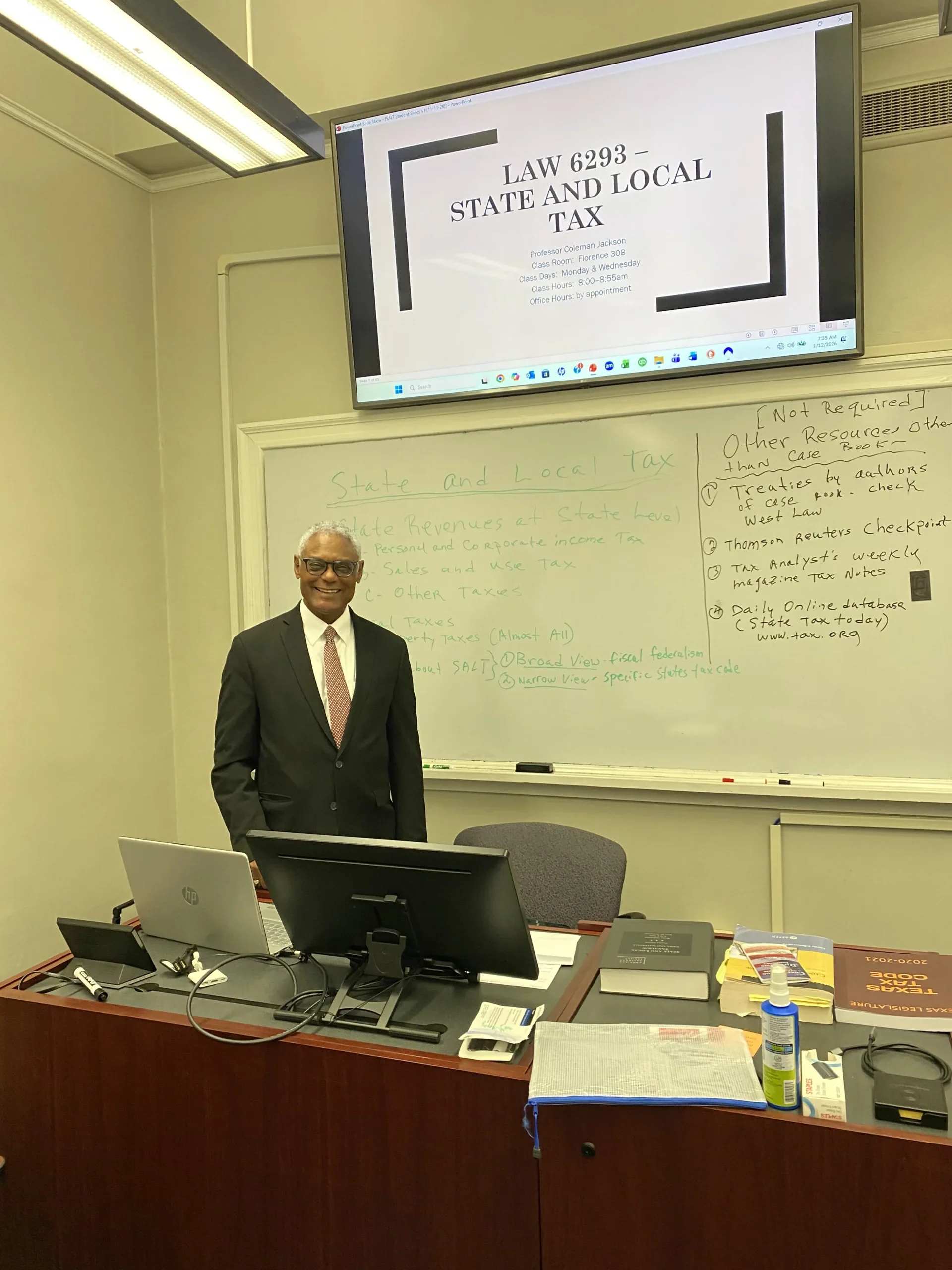 Professor standing in a classroom with a whiteboard and screen displaying "Law 6293 - State and Local Tax.