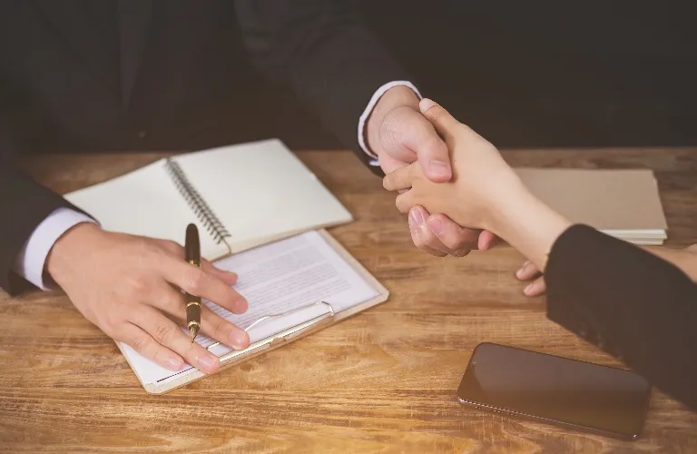 Two individuals in business attire shaking hands over a wooden desk with documents, a pen, a notebook, and a smartphone.
