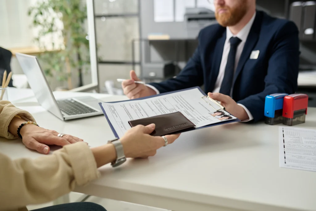 Two individuals exchanging a clipboard with documents and passport in a professional office setting.