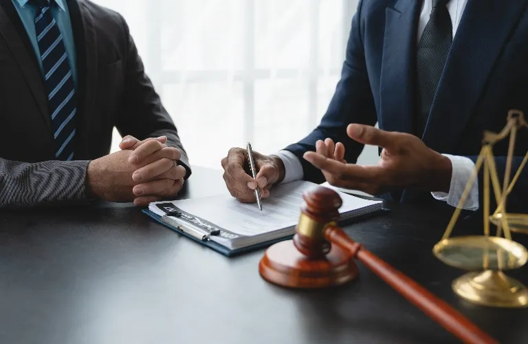 Two men in suits discussing legal documents with gavel and scales of justice on the table.