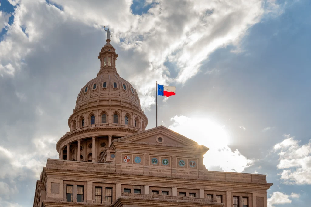 Texas State Capitol dome with Texas flag against partly cloudy sky and sun rays.