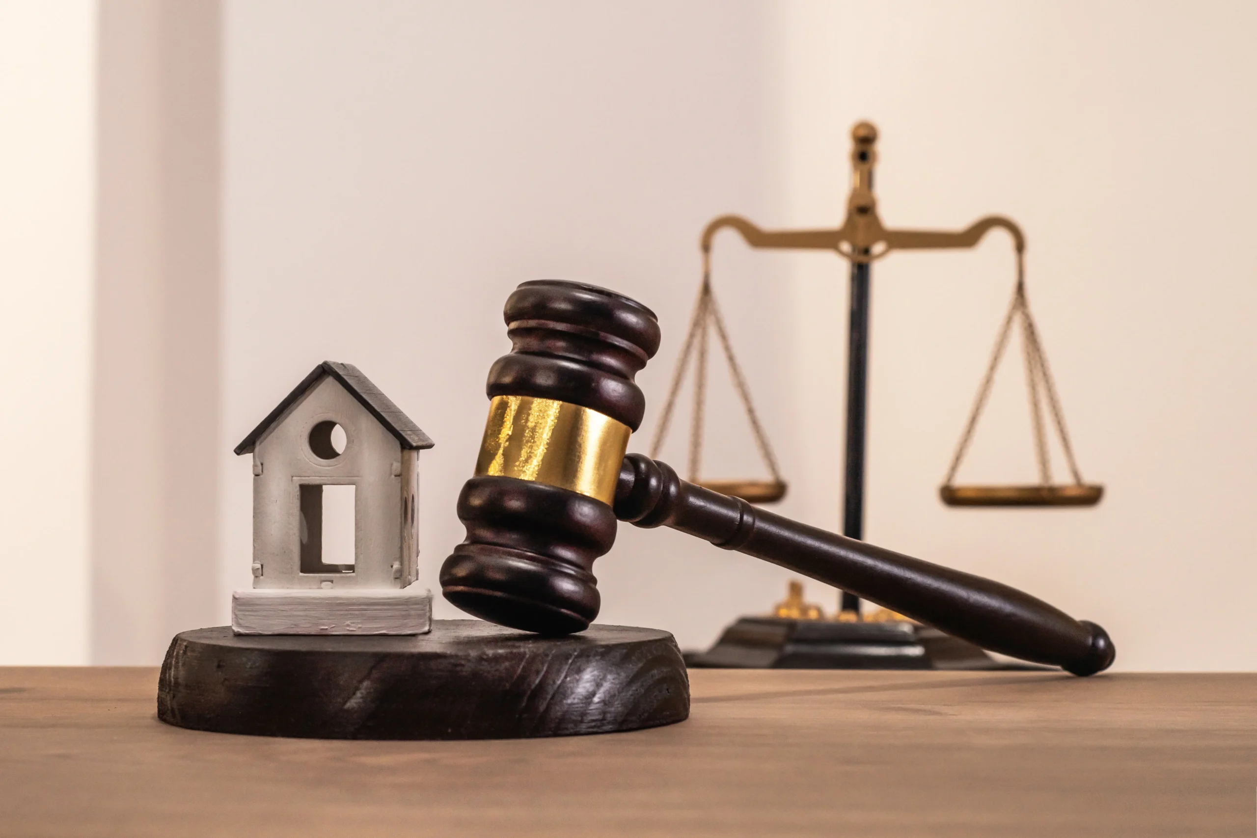 Wooden judge's gavel with house model and brass balance scales on desk.