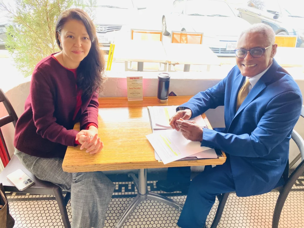Two professionally dressed individuals seated at a wooden table with documents and a coffee tumbler.