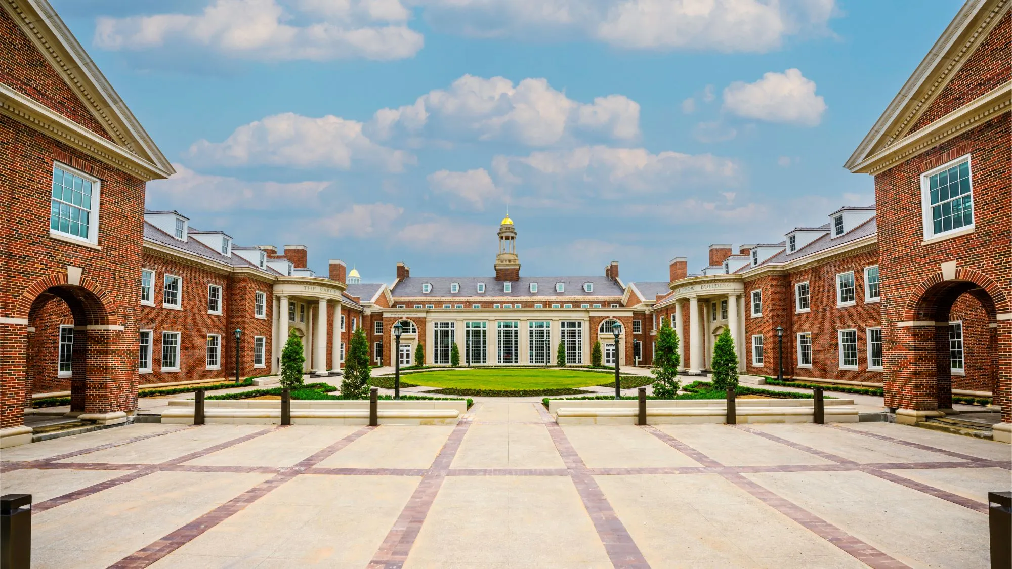 Symmetrical red brick academic buildings with white columns surrounding a green courtyard under a blue sky.