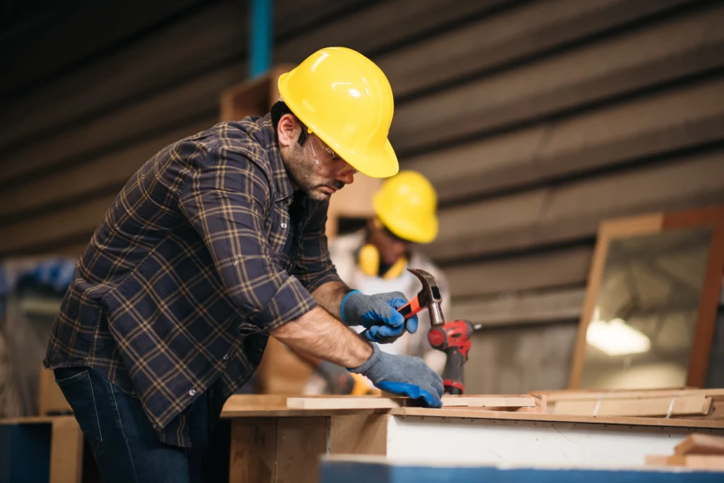 Two construction workers wearing yellow hard hats and safety glasses working with wood and tools indoors.