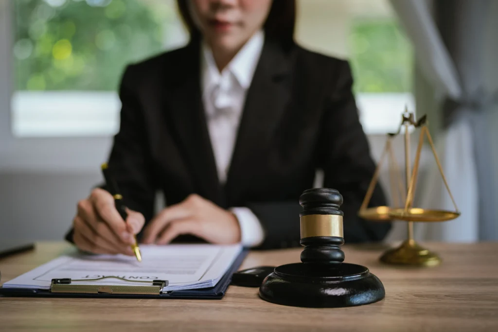 Person in formal black suit signing a contract at a desk with a judge's gavel and scales of justice.