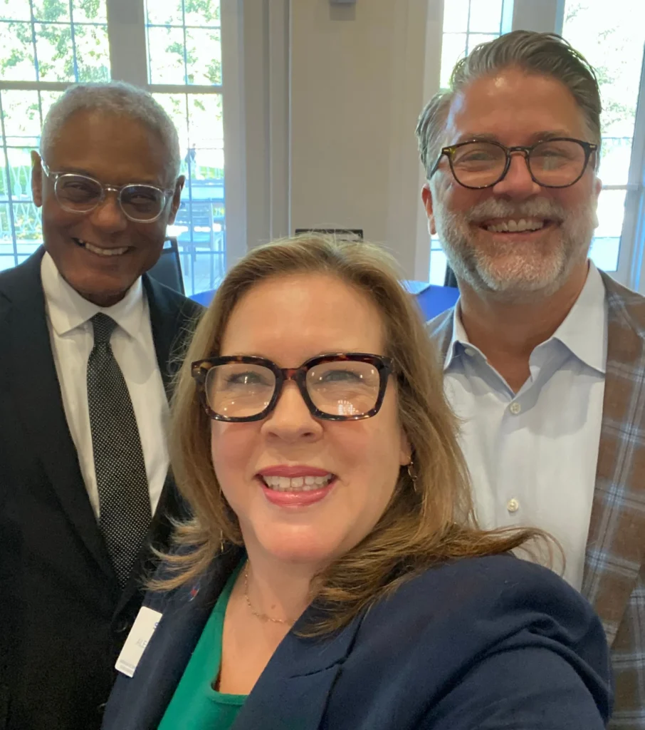 Three professionally dressed individuals smiling, standing indoors with large windows in the background.