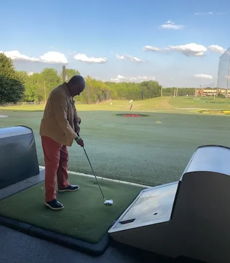 Man in beige jacket and red pants preparing to hit golf ball at driving range.