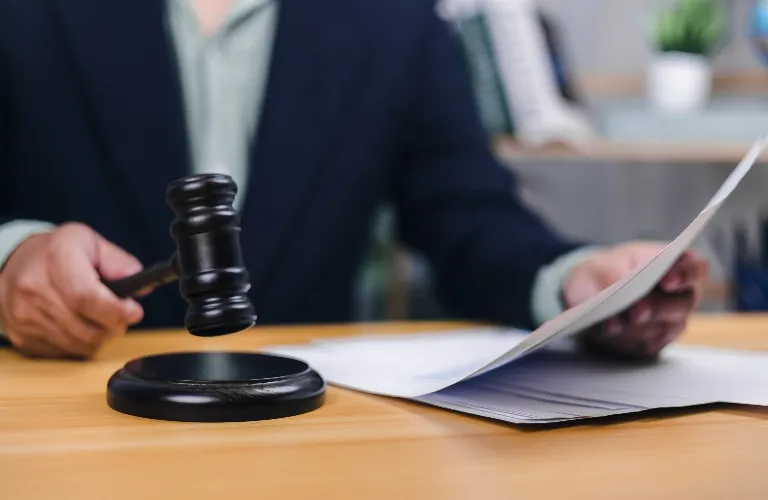 Person in dark suit holding wooden gavel and reviewing documents on desk.