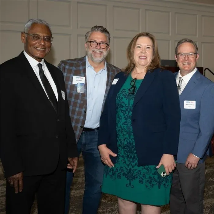 Four professionally dressed individuals standing indoors against a beige paneled wall, smiling at the camera.