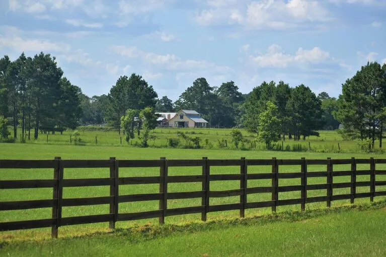 Rural landscape featuring a wooden fence, green pasture, scattered trees, and a distant farmhouse under a partly cloudy sky.
