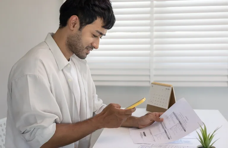 Man in white shirt using smartphone to scan documents at white desk with calendar and plant.