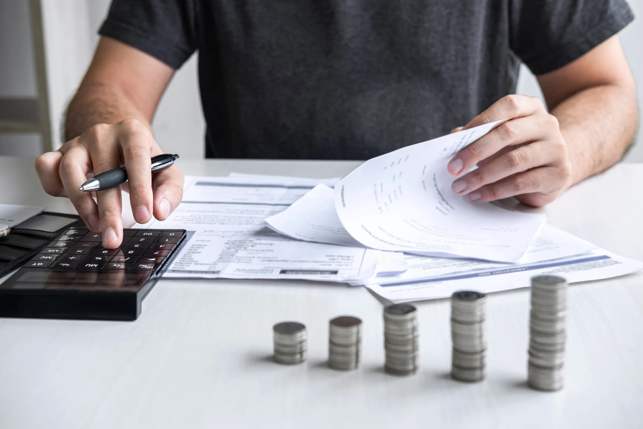 Person using calculator and reviewing documents with stacked coins on desk.