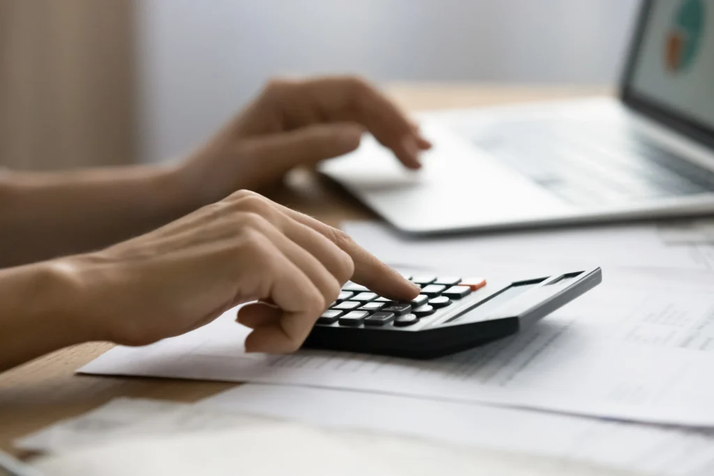 Close-up of hands using a calculator and laptop on a desk with documents.