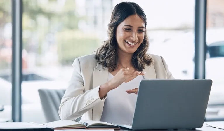 Professional woman in beige blazer smiling and gesturing during video call on laptop.