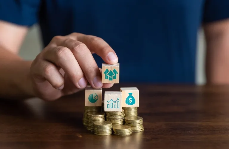 Hand placing wooden block with upward arrows icon on stacks of gold coins and financial symbol blocks.