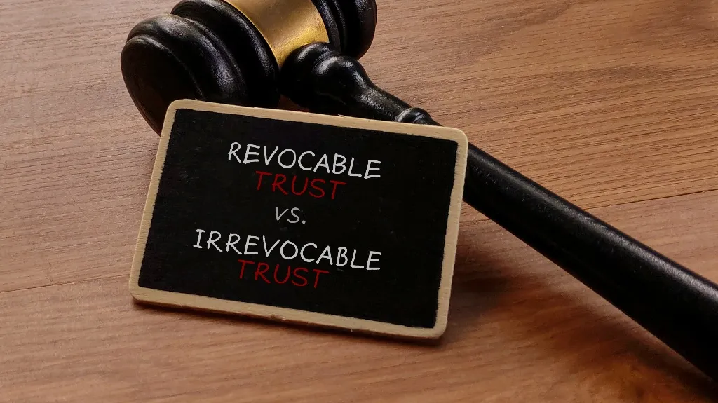 Wooden judge's gavel resting on a table next to a small blackboard with "Revocable Trust vs. Irrevocable Trust" text.