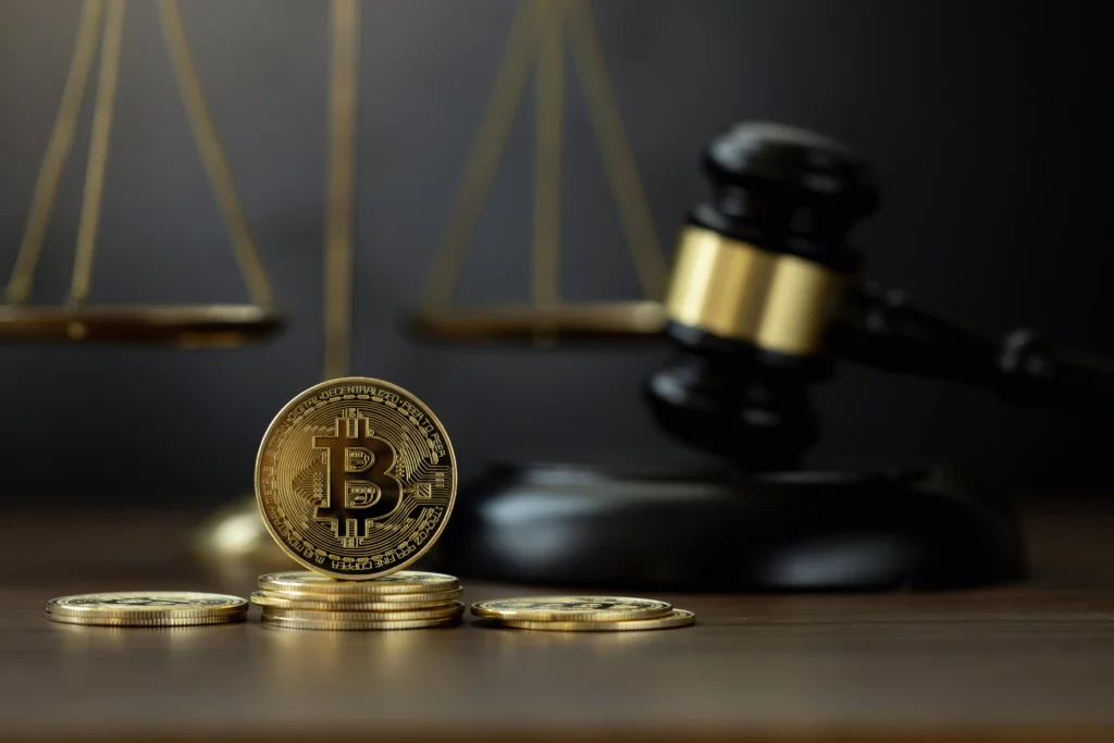 Stack of gold Bitcoin coins in foreground with blurred scales of justice and judge's gavel in background.