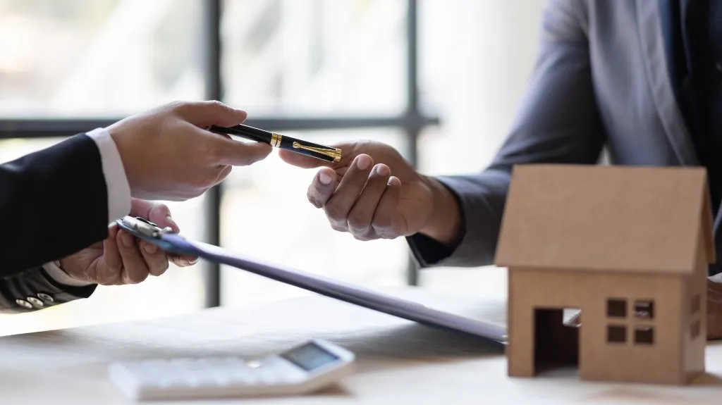 Two individuals exchanging a pen over a clipboard near a small wooden house model on a table.
