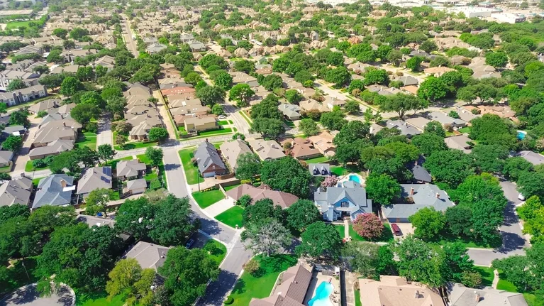 Aerial view of a suburban neighborhood with numerous houses, green trees, and swimming pools.