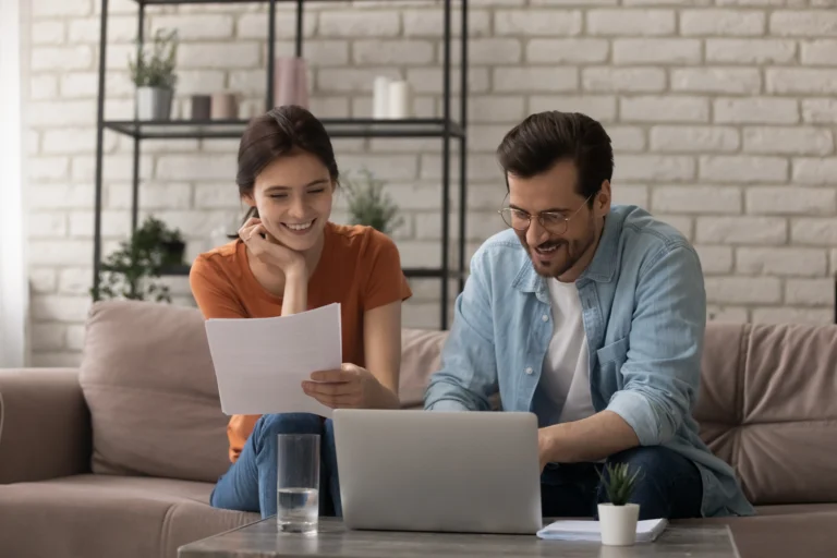 Young woman and man smiling while reviewing documents and using laptop on beige couch.