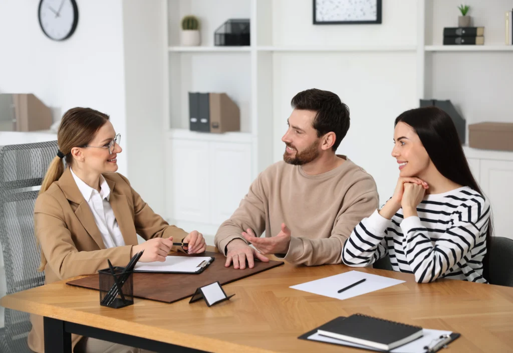 Professional woman in beige suit consulting smiling couple in office with white shelves and clock.