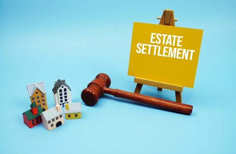 Wooden gavel, miniature houses, and yellow sign on easel reading "Estate Settlement" on blue background.