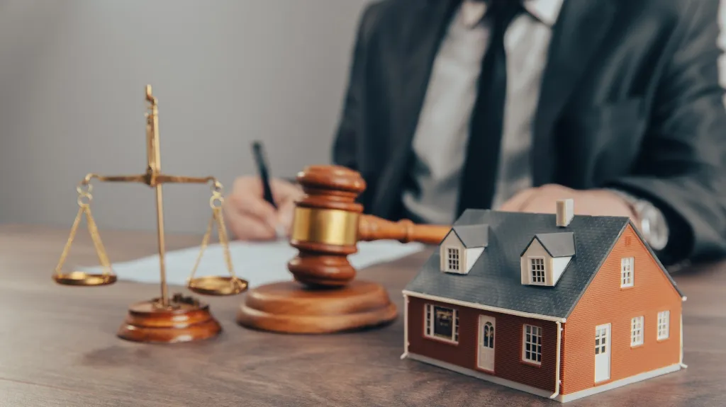 Close-up of a wooden gavel, gold scales of justice, and a model house on a desk with a person in a suit writing in the background.
